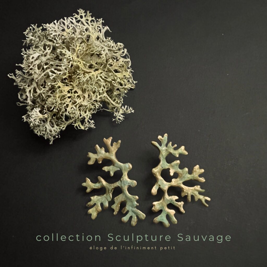 Paire de boucles d'oreilles en bronze d'art patiné inspirée de la forme du lichen Mousse de chêne, posée à côté d'un véritable lichen sur un fond noir, Collection Sculpture Sauvage de l'atelier Herbarium.