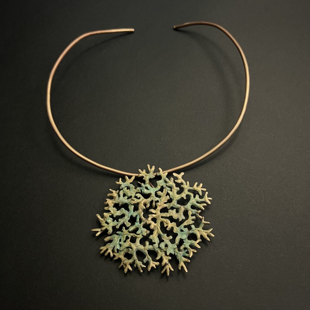 Un bijou en bronze d'art modulable de la collection Sculpture Sauvage par l'atelier Herbarium (Jane Bosshard Quillerat). Un collier torque rigide et ouvert en bronze doré soutient un grand pendentif circulaire et ajouré. Ce médaillon reproduit fidèlement la texture organique et ramifiée du lichen mousse de chêne, mêlant des reflets dorés à une patine texturée aux nuances vert-de-gris. Le bijou est photographié de dessus sur un fond noir uni.