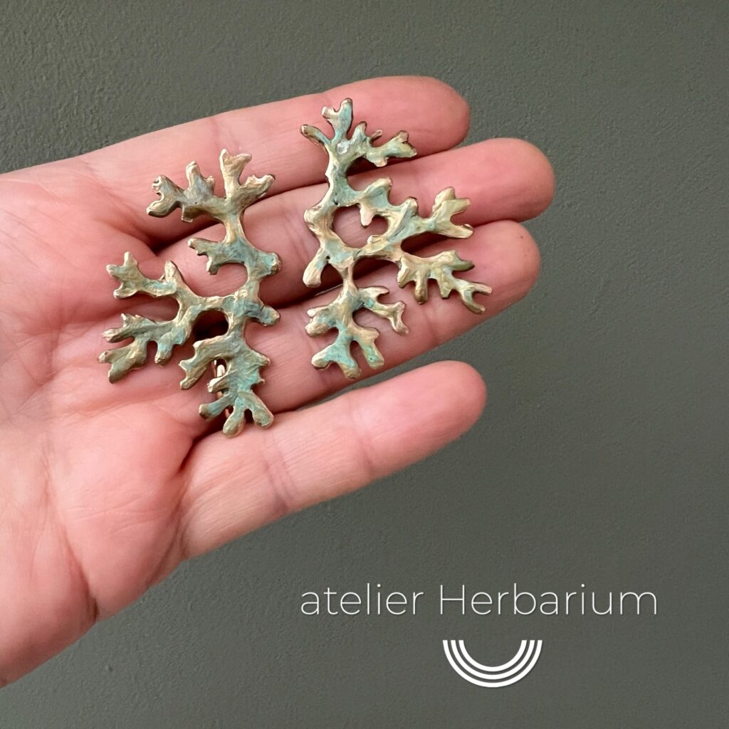Paire de boucles d'oreilles en bronze d'art patiné inspirée de la forme du lichen Mousse de chêne, présentée dans un main. photographiée sur fond vert de gris. Collection Sculpture Sauvage de l'atelier Herbarium.