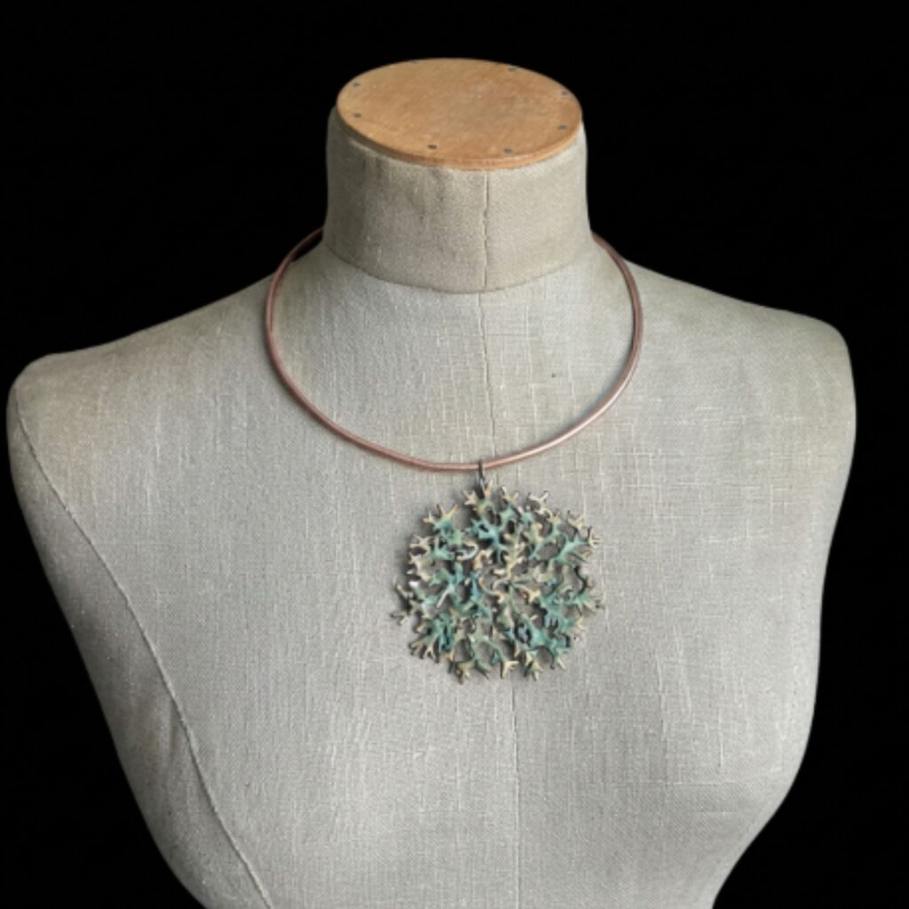 Un bijou en bronze d'art modulable de la collection Sculpture Sauvage par l'atelier Herbarium (Jane Bosshard Quillerat). Un collier torque rigide et ouvert en bronze doré soutient un grand pendentif circulaire et ajouré. Ce médaillon reproduit fidèlement la texture organique et ramifiée du lichen mousse de chêne, mêlant des reflets dorés à une patine texturée aux nuances vert-de-gris. Le bijou est photographié sur un buste en lin sur fond noir.