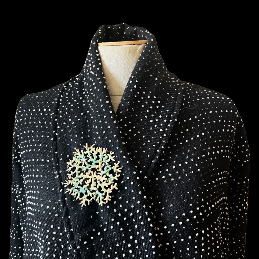 Le bijou modulable en bronze d'art de l'atelier Herbarium est ici porté en broche sur un mannequin de couture. Le grand médaillon circulaire, sculpté en forme de lichen mousse de chêne aux nuances dorées et vert-de-gris, est épinglé sur le revers croisé d'un manteau noir texturé à motifs de petits points blancs. L'ensemble est présenté sur un fond noir uni.