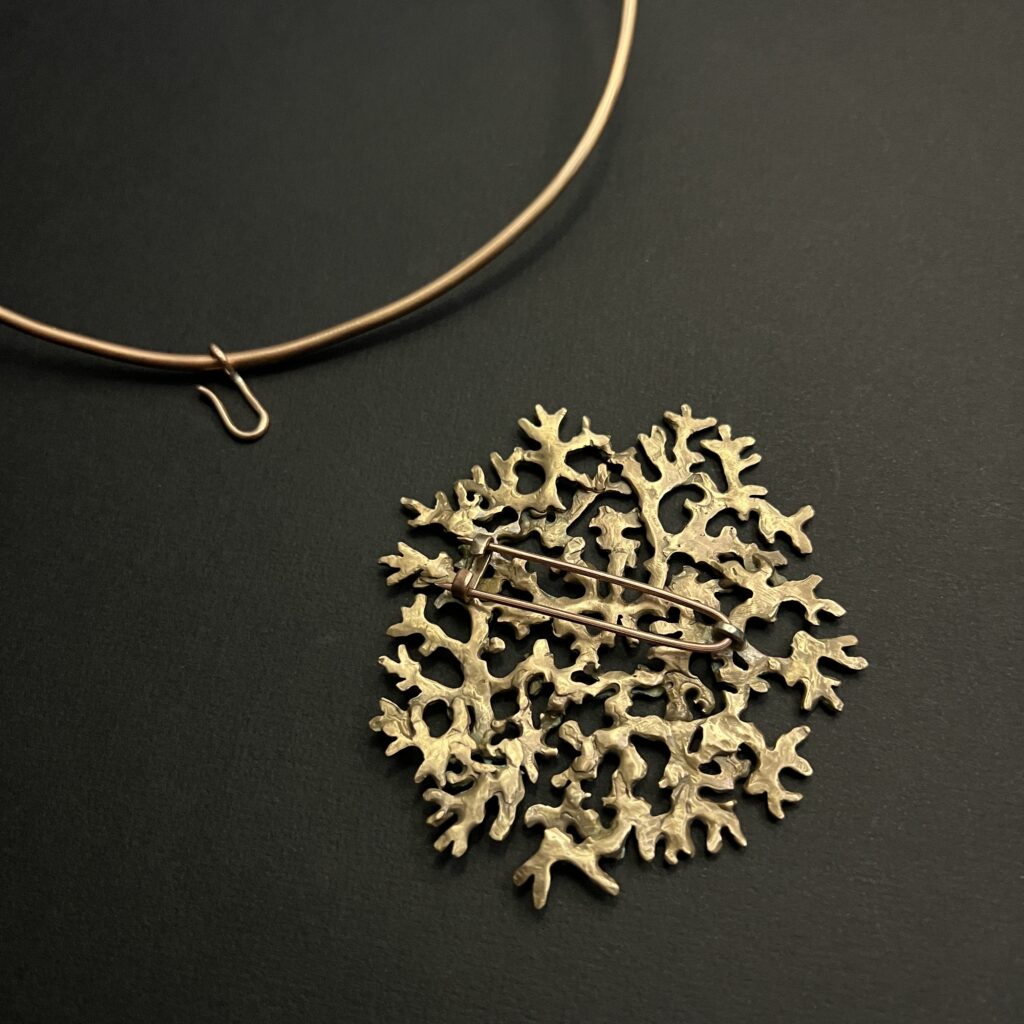 Verso du bijou en bronze d'art modulable de la collection Sculpture Sauvage par l'atelier Herbarium (Jane Bosshard Quillerat). Un collier torque rigide et ouvert en bronze doré soutient un grand pendentif circulaire et ajouré. Ce médaillon reproduit fidèlement la texture organique et ramifiée du lichen mousse de chêne, mêlant des reflets dorés à une patine texturée aux nuances vert-de-gris. Le bijou est photographié de dessus sur un fond noir uni.