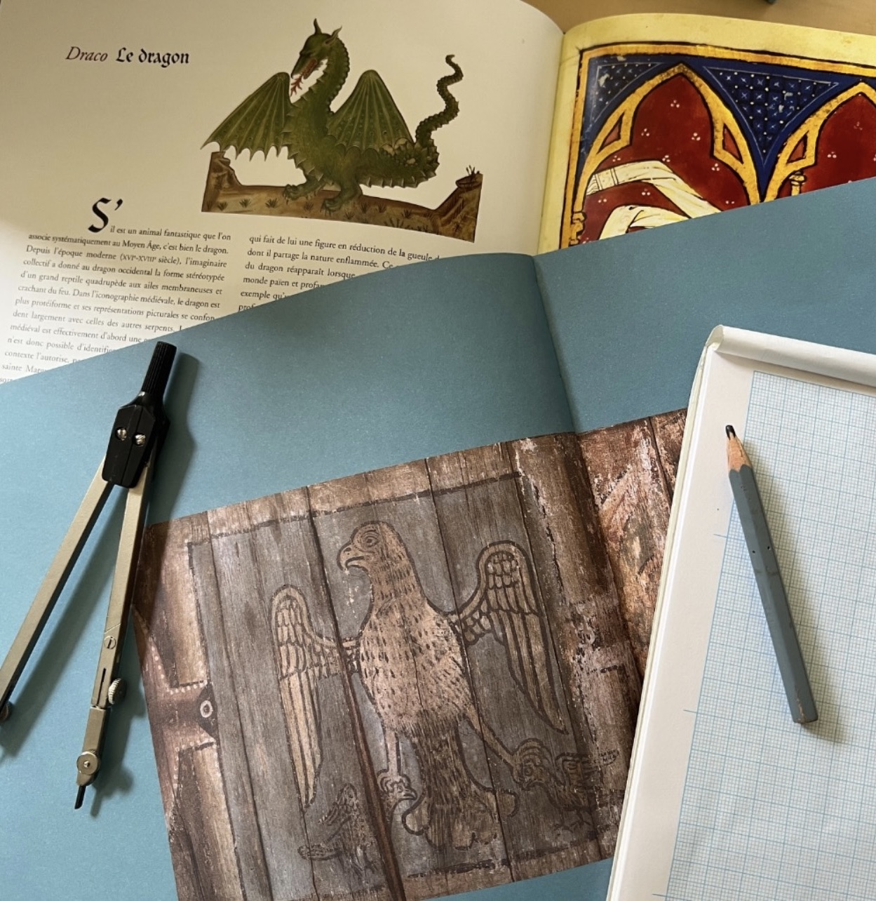 Outils de dessin sur l'établi avec un compas, un crayon et du papier à côté de livres ouverts sur des bestiaires médiévaux pour les croquis d'une nouvelle collection de bijoux.
